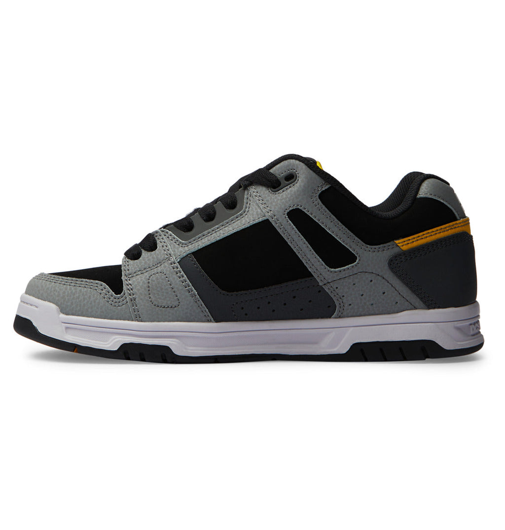 The Men’s Stag Shoes by DC Shoes are athletic sneakers in gray and black, featuring a white sole with a sturdy cupsole, subtle yellow accents, and are displayed in a side view against a white background.