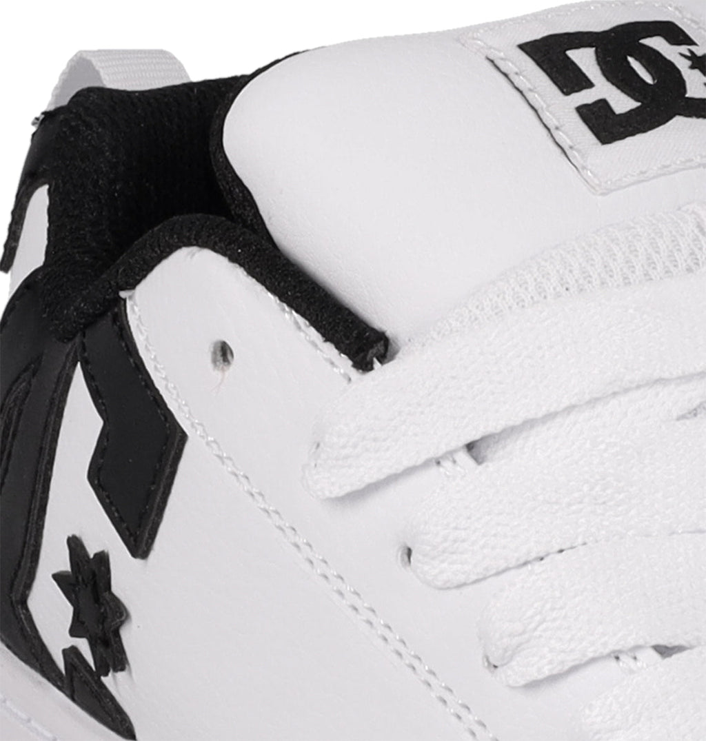 Close-up of DC Shoes Kids' Court Graffik: a white sneaker with black accents, visible stitching, white laces, black inner lining, and DC logos by the laces and heel. Skate-inspired style from DC Shoes.