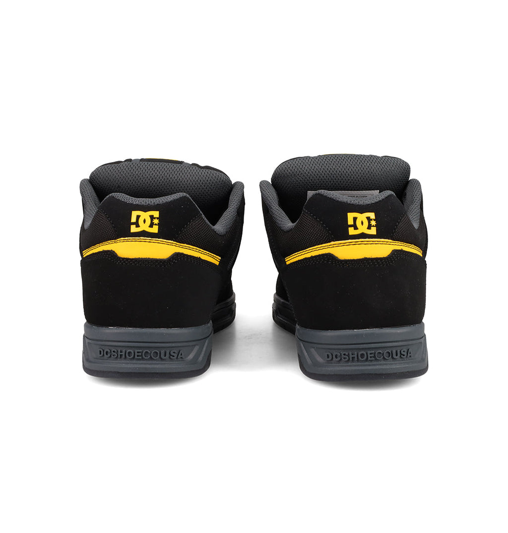 The Men's Stag Shoes by DC Shoes in black with yellow accents and logo detail on the heels feature a durable cupsole, shown from the back on a white background—ideal for authentic skate culture.