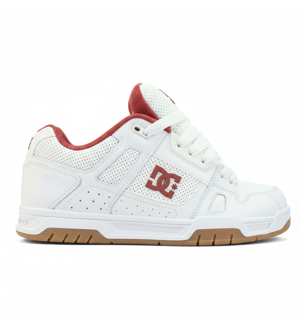 The Men’s Stag Shoes by DC Shoes are white skate shoes featuring red accents, a DC logo on the side, red lining, perforated details, an oversized puff tongue, thick white laces, and a durable cupsole gum rubber sole. The shoes are shown in a side profile view against a white background.