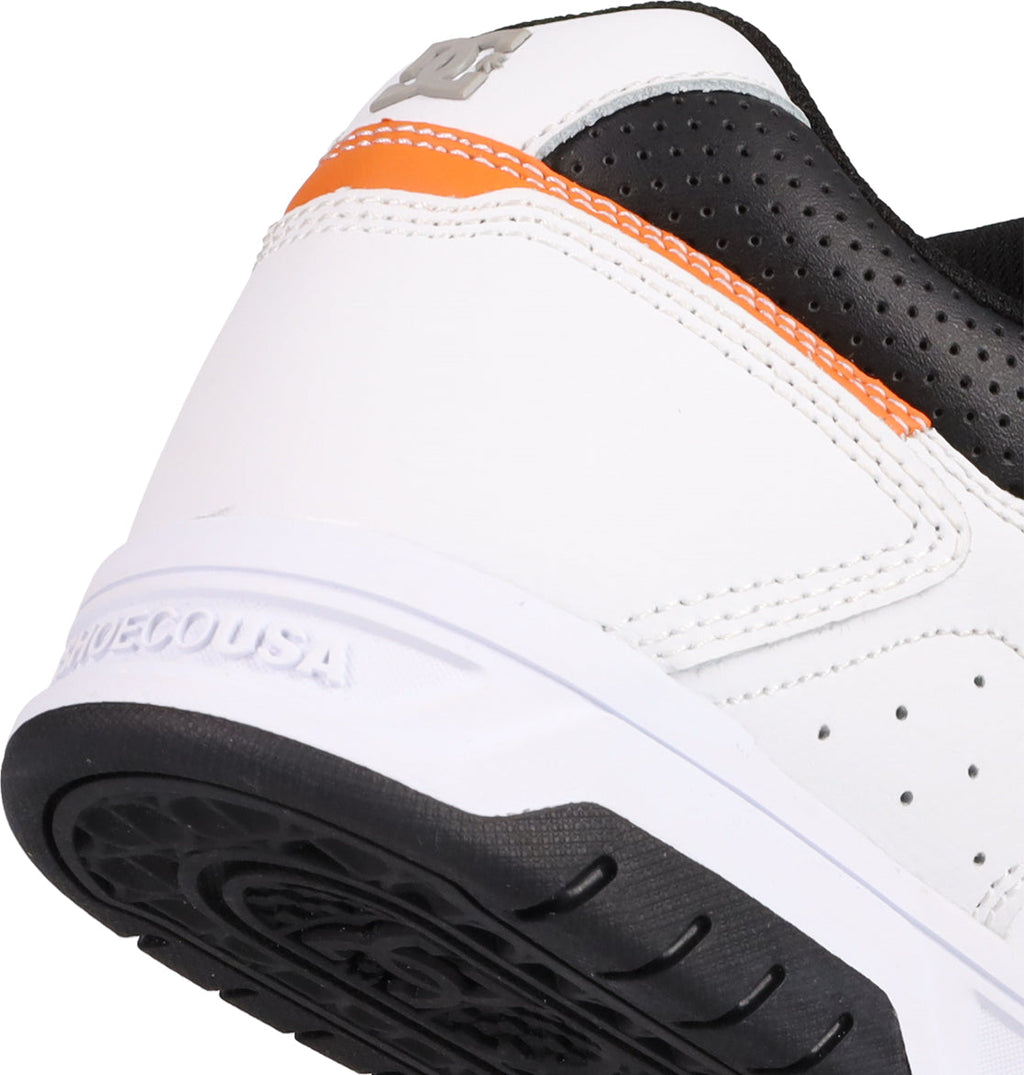 Close-up of the heel of a white Men's Stag Shoe by DC Shoes, featuring black and orange details, a textured cupsole, visible stitching, an embossed brand logo on the heel, and SHOECOUSA molded into the rubber sole—a nod to skate culture.