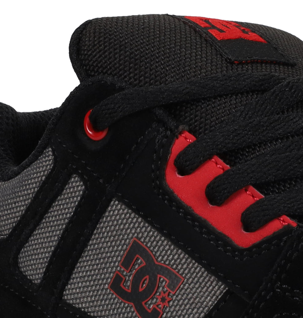 Close-up of a black and gray Kids’ Stag Shoe by DC Shoes with red accents, featuring the brand logo, laces, and detailed textured fabric. The sneaker offers lightweight cushioning for comfort and rubber elements engineered to enhance impact absorption.
