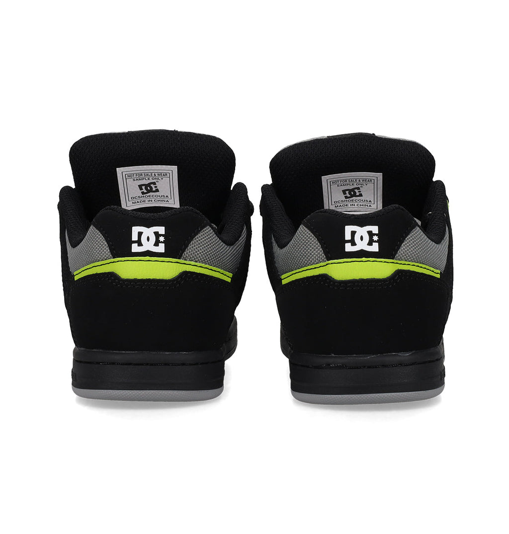 Back view of a pair of Kids’ Stag Shoes by DC Shoes in black with gray accents and a neon yellow stripe, showcasing the iconic DC logo on the heels and inner labels. This minimal silhouette offers everyday versatility as a go-to vulc shoe for effortless style and comfort.