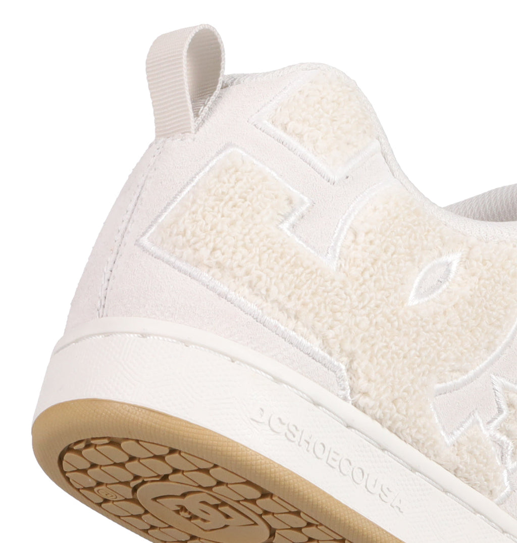 Close-up of the heel and side of DC Shoes Women's Court Graffik SD, featuring a white suede upper, textured fuzzy lettering, beige rubber sole, pull tab at the back, visible stitching details, and fashion-forward colorways.