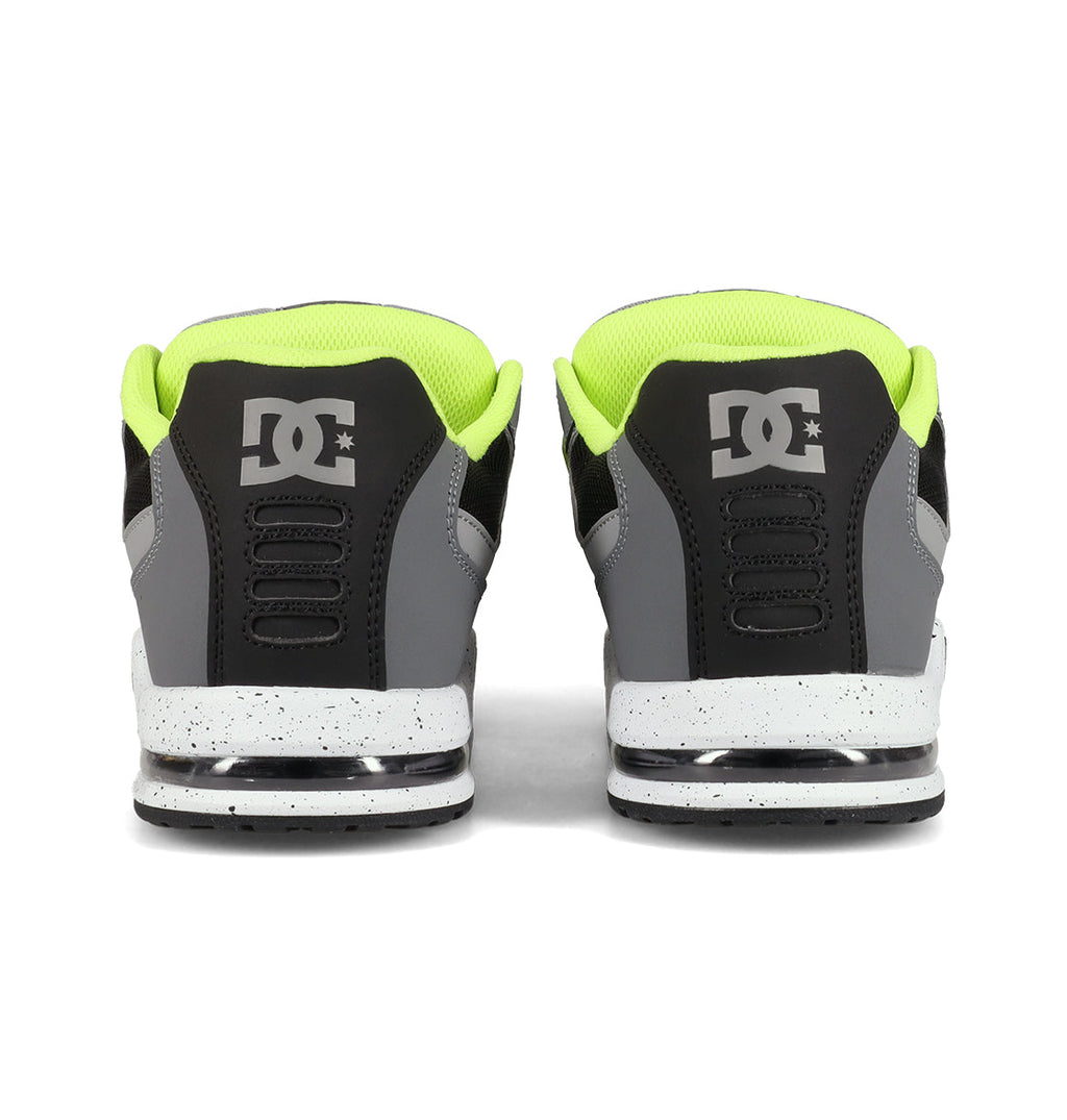 A pair of DC Shoes Men’s AT-2 Shoes is shown from the back, showcasing a black and gray color scheme accented by neon yellow lining and white speckled soles. The DC logo appears on each heel, while superior cushioning delivers lasting comfort with every step.