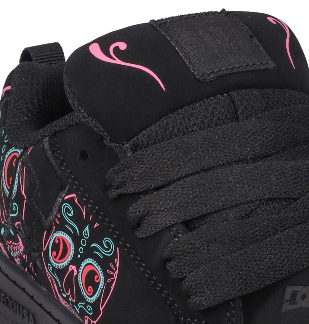 Close-up of the Kids’ Court Graffik Day Of The Dead sneaker by DC Shoes, showcasing thick black laces and vibrant pink and teal skull designs inspired by Día de los Muertos on the side and near the tongue. The black sneaker features a fabric pull tab and a plain black patch on the tongue.