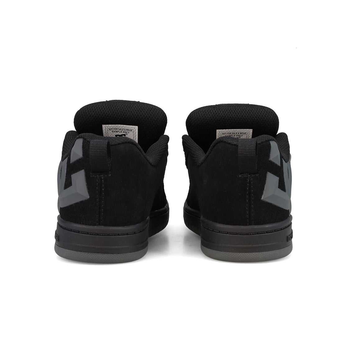A pair of Kids’ Court Graffik Beveled Special Edition Shoes by DC Shoes are shown from the back, featuring thick black soles and a dark geometric beveled TPR rubber logo on each heel for signature DC branding. The black low-top sneakers are positioned side by side against a white background.