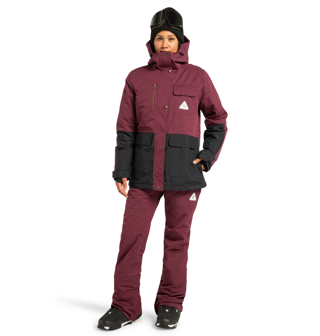 Wearing the DC Shoes Women’s Liberate Insulated Jacket in maroon and black, paired with matching maroon snow pants, black gloves, black boots, and a black helmet with goggles, this person stands against a plain white background—perfectly outfitted for any winter adventure with reliable weather protection.