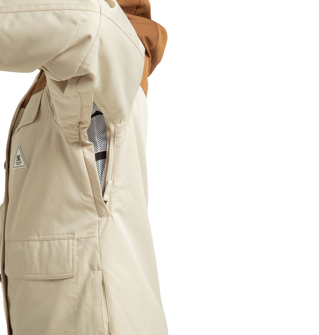 A close-up of a beige DC Shoes Women’s Liberate Insulated Jacket with a brown hood highlights an unzipped side vent, revealing the mesh lining inside. Crafted from recycled polyester, this jacket is ideal for winter adventures. The wearer has their arm raised, showing a flap pocket on the side.