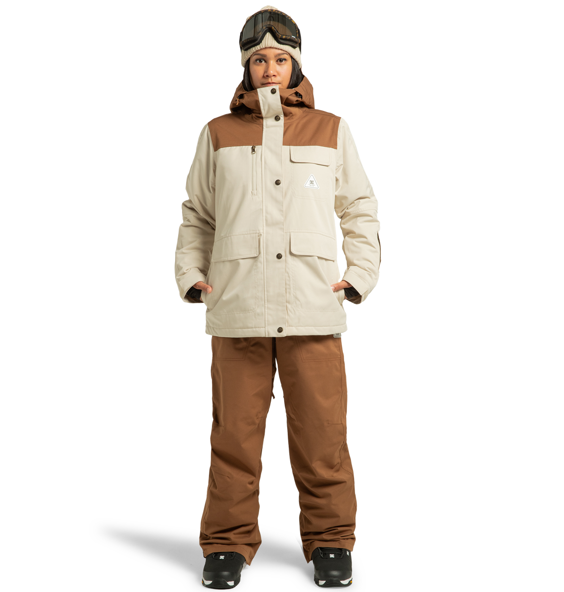 A person stands against a plain white background, ready for a winter adventure in the DC Shoes Women’s Liberate Insulated Jacket and matching beige and brown pants made from recycled polyester, complemented by black gloves, black boots, and ski goggles resting on their head.