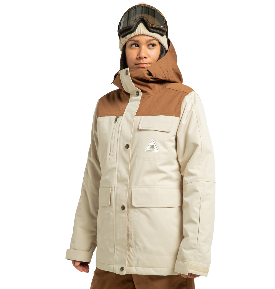 A person models the DC Shoes Women’s Liberate Insulated Jacket in beige and brown, crafted from recycled polyester, paired with brown pants and ski goggles atop a knit hat, gazing to the side against a plain white background—ready for any winter adventure.