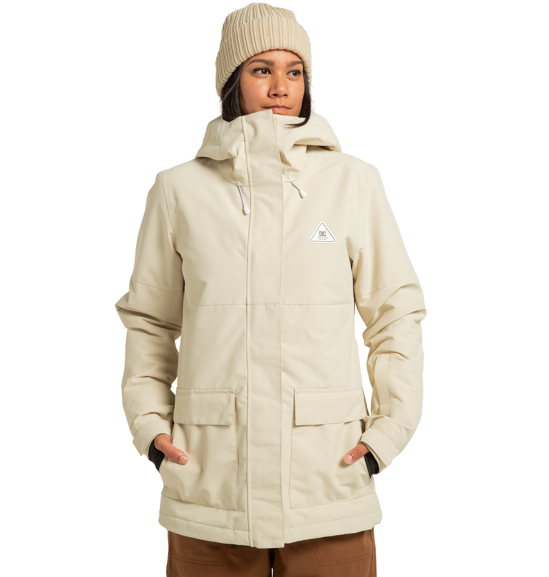 A person stands against a plain white background with hands in the pockets of the DC Shoes Women’s Cruiser Insulated Jacket—a beige, hooded winter jacket paired with a matching knit beanie. Made from recycled polyester and enhanced with weather defense technology.