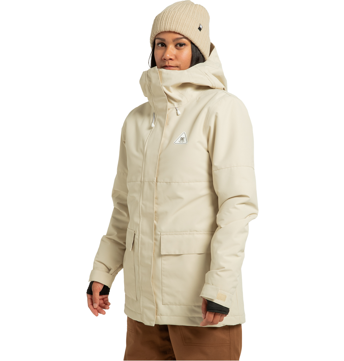 A woman stands against a plain white background, facing slightly to the side with a neutral expression, wearing the DC Shoes Women’s Cruiser Insulated Jacket in beige—crafted from recycled polyester and featuring weather defense technology—paired with brown pants and a beige knit beanie.