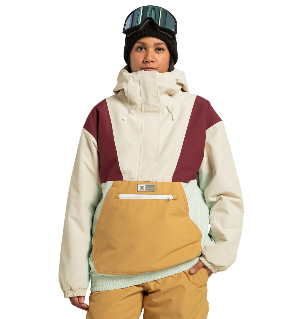 A person stands against a plain white background, ready for any outdoor adventure, wearing the DC Shoes Women’s Chalet Anorak Insulated Jacket—crafted from recycled polyester with beige, maroon, mint, and mustard colorblocking—paired with tan pants, a black beanie, and ski goggles.