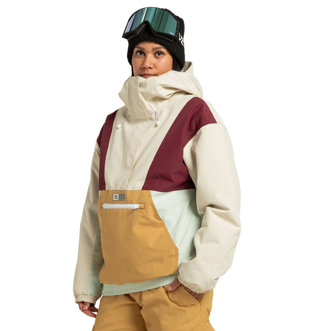A person stands against a plain white background wearing the DC Shoes Women’s Chalet Anorak Insulated Jacket—crafted from recycled polyester with beige, maroon, light blue, and mustard panels—paired with tan snow pants, a black beanie, and ski goggles.