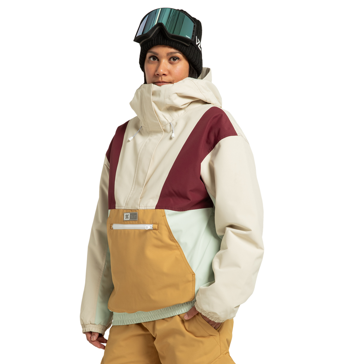 A person stands against a plain white background wearing the DC Shoes Women’s Chalet Anorak Insulated Jacket—crafted from recycled polyester with beige, maroon, light blue, and mustard panels—paired with tan snow pants, a black beanie, and ski goggles.