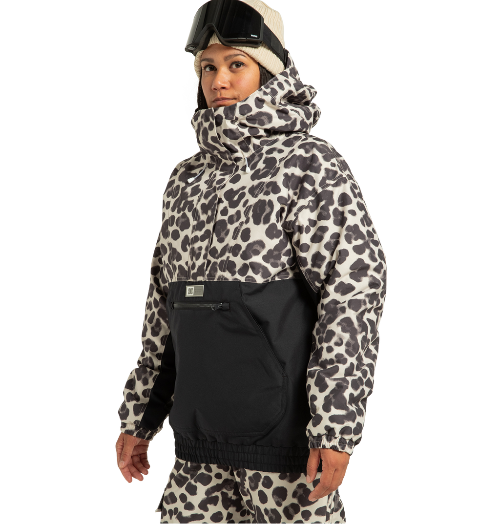 A person wearing a beige beanie, ski goggles, and the DC Shoes Women’s Chalet Anorak Insulated Jacket with a leopard print design and black accents stands facing slightly left against a plain background. This stylish women’s jacket features recycled polyester for an eco-friendly touch.