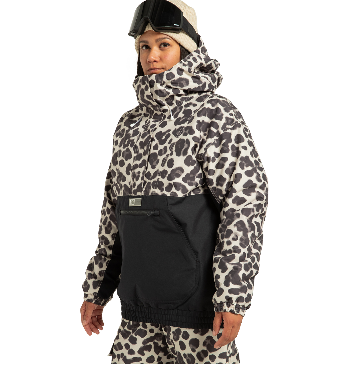 A person wearing a beige beanie, ski goggles, and the DC Shoes Women’s Chalet Anorak Insulated Jacket with a leopard print design and black accents stands facing slightly left against a plain background. This stylish women’s jacket features recycled polyester for an eco-friendly touch.