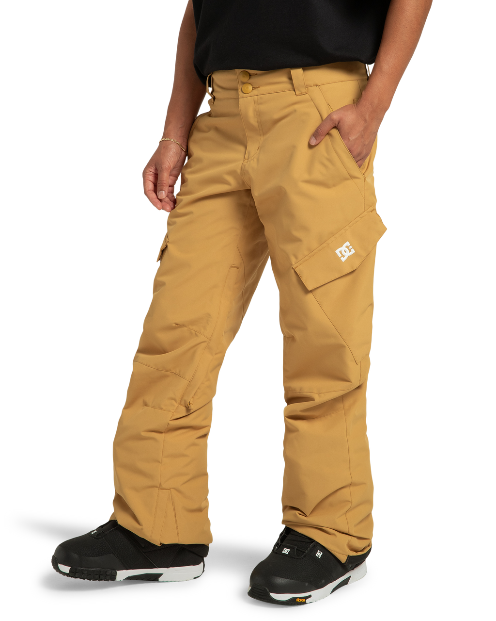 A woman stands against a white background wearing DC Shoes Women’s Nonchalant Insulated Snow Pants in tan, featuring the DC Shoes logo on the pocket. She pairs them with a black shirt and black sneakers. These snow pants are made from recycled polyester, combining style with sustainability.