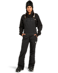 A person wearing the DC Shoes Women’s Crusade Shell Bib in black, paired with a black sweatshirt, black boots, beanie, and ski goggles—standing with hands in pockets against a white background—showcases women’s adventure gear engineered for ultimate waterproof protection.