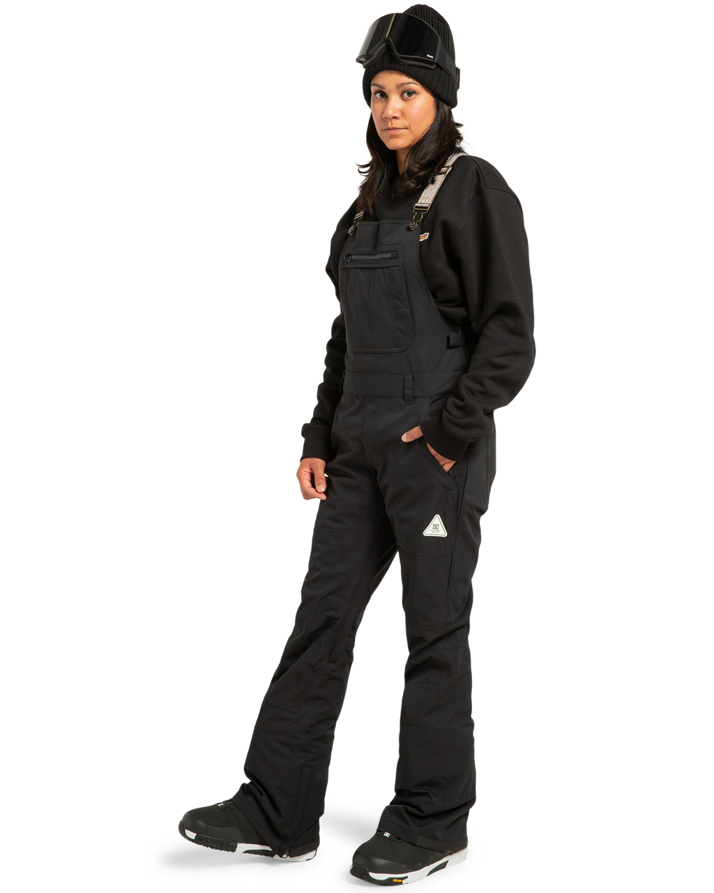 A person stands facing forward against a white background, wearing DC Shoes Women’s Crusade Shell Bib black snow overalls, paired with a black sweatshirt, black boots, a black beanie, and ski goggles; their arms are relaxed by their sides as they showcase waterproof women’s adventure gear.