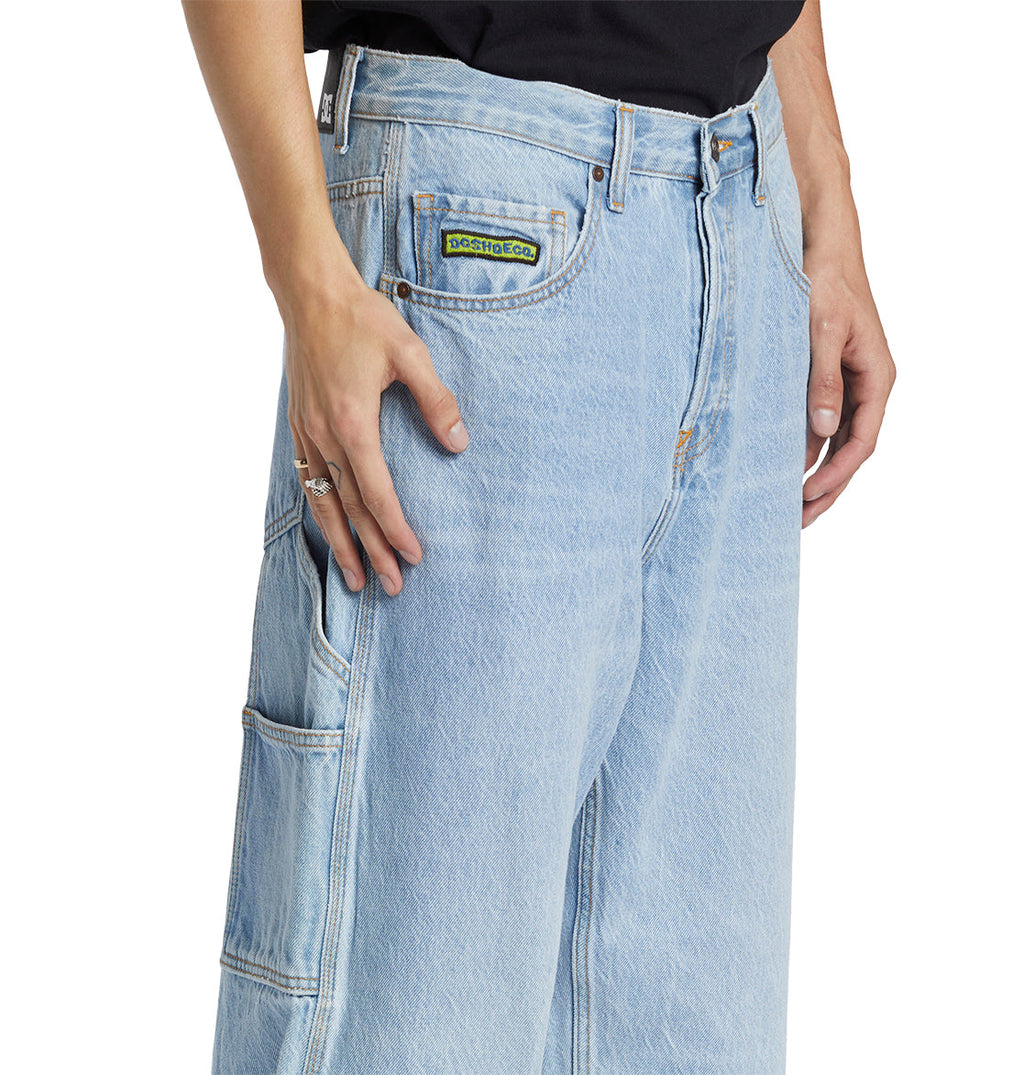 A person is shown from the waist to mid-thigh wearing DC Shoes Men’s Worker Baggy Carpenter Ril Jeans in light blue. The jeans feature a relaxed, loose fit with classic workwear-inspired seam detailing down the leg and a distinctive green and yellow patch near the pocket. One hand rests casually on the thigh.