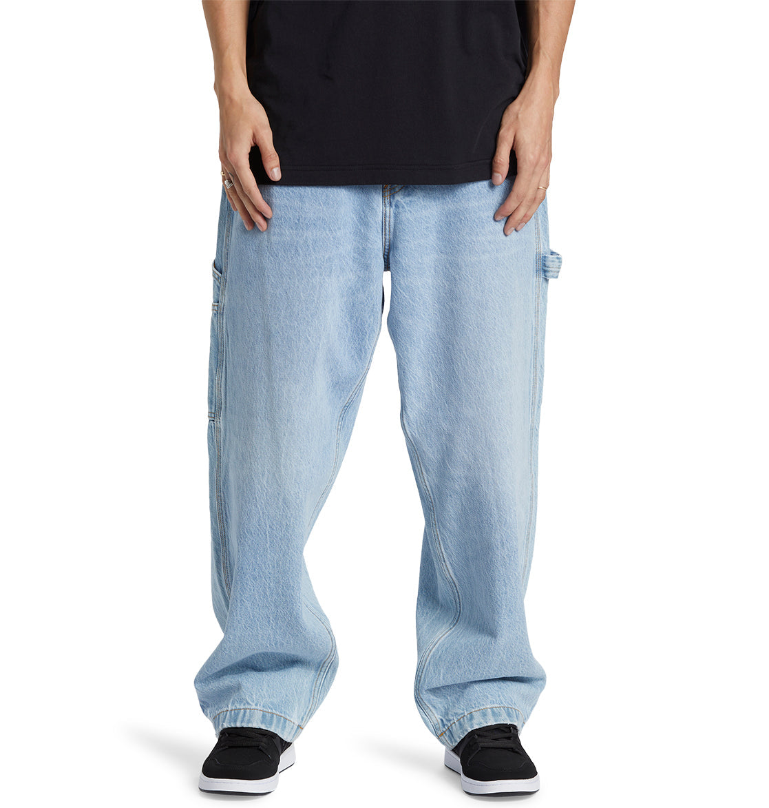 A person is wearing DC Shoes Men’s Worker Baggy Carpenter Ril Jeans in light blue, paired with a black shirt and black sneakers, standing with arms relaxed by their sides. The photo is cropped from the shoulders down.