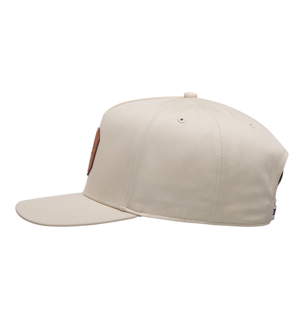 The Men's Reynotts Cap by DC Shoes is displayed side-on against a white background. This beige skate cap features a round brown DC patch on the front and visible stitching along its panels.