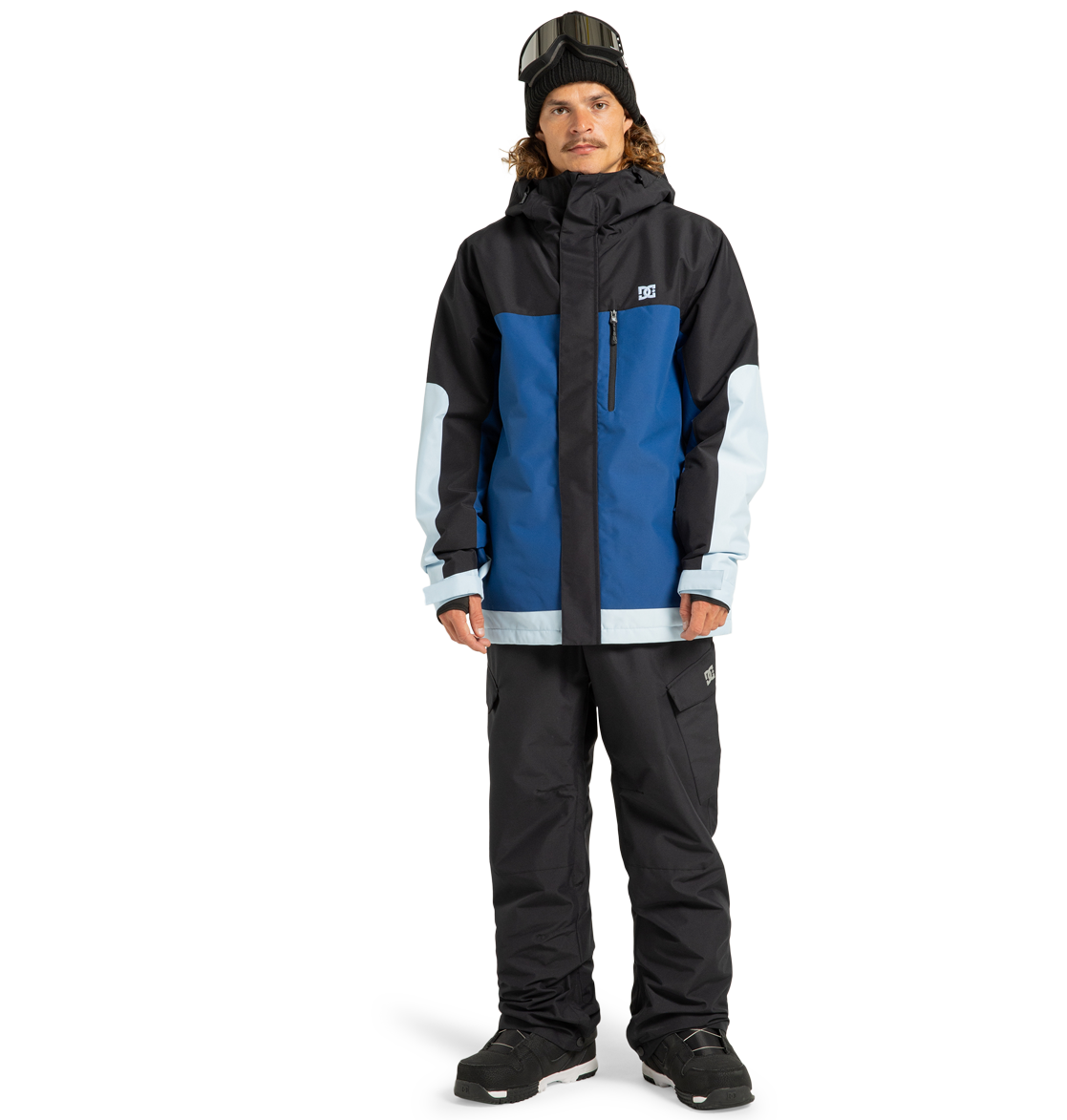 A person wearing the DC Shoes Men’s Defiant Insulated Jacket—featuring a colorblock design in blue, black, and light blue with recycled polyester construction and Weather Defense 10 technology—is paired with black snow pants, black boots, a black beanie, and ski goggles while standing against a plain white background.
