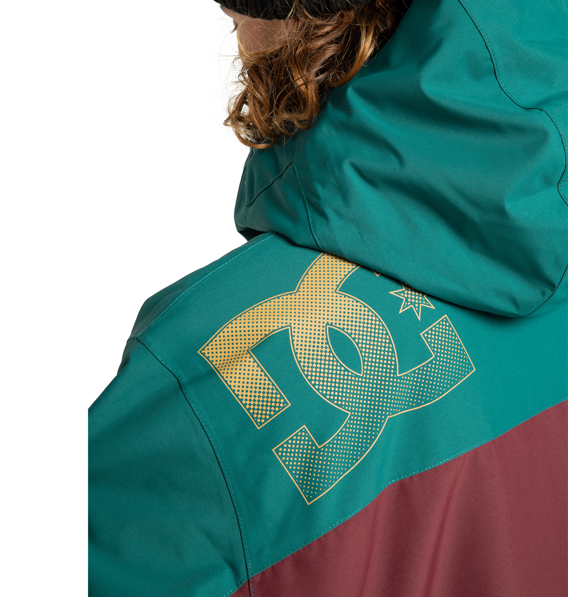A person with brown curly hair wears the DC Shoes Men’s Defiant Insulated Jacket in teal and maroon, featuring a large yellow DC logo on the back shoulder. The hood is up, partially covering their head.