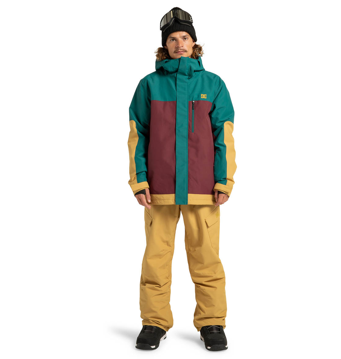 A person wearing the DC Shoes Men’s Defiant Insulated Jacket in teal, burgundy, and tan, styled with tan snow pants, black gloves, black boots, and ski goggles on their head stands against a plain white background.