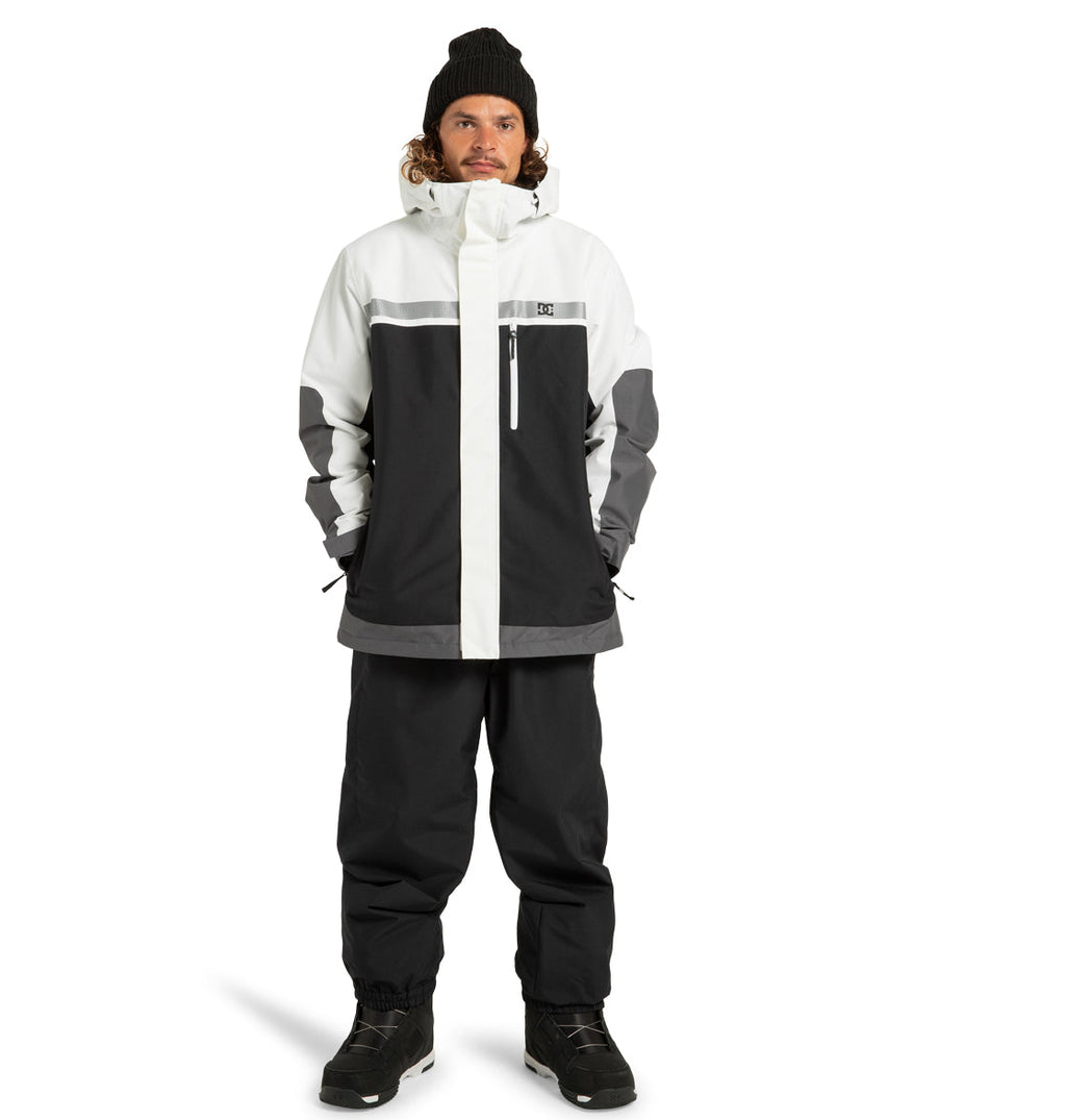 A man wearing the DC Shoes Men’s Defiant Insulated Jacket in black and white—crafted from recycled polyester—paired with black snow pants, black boots, and a black knit hat, stands against a plain white background.