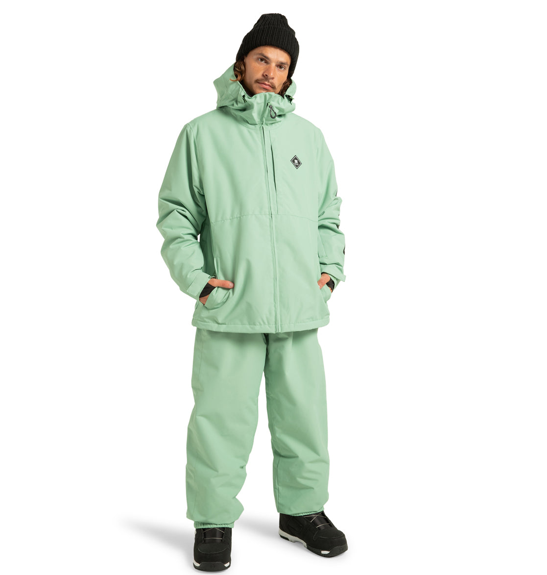 A person wearing the DC Shoes Unisex Basis Insulated Jacket in mint green, paired with matching pants, stands with hands in pockets. Made from recycled polyester, this outfit is perfect for winter pursuits. They complete the look with a black beanie and black shoes while posing against a plain white background.