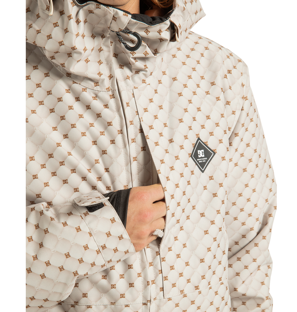 A person wearing the DC Shoes Unisex Basis Print Insulated Jacket, a light-colored patterned outerwear piece with a diamond-shaped logo on the chest, partially unzipped to reveal a black inner layer. Crafted from recycled polyester, this jacket features a high collar and adjustable cuffs.