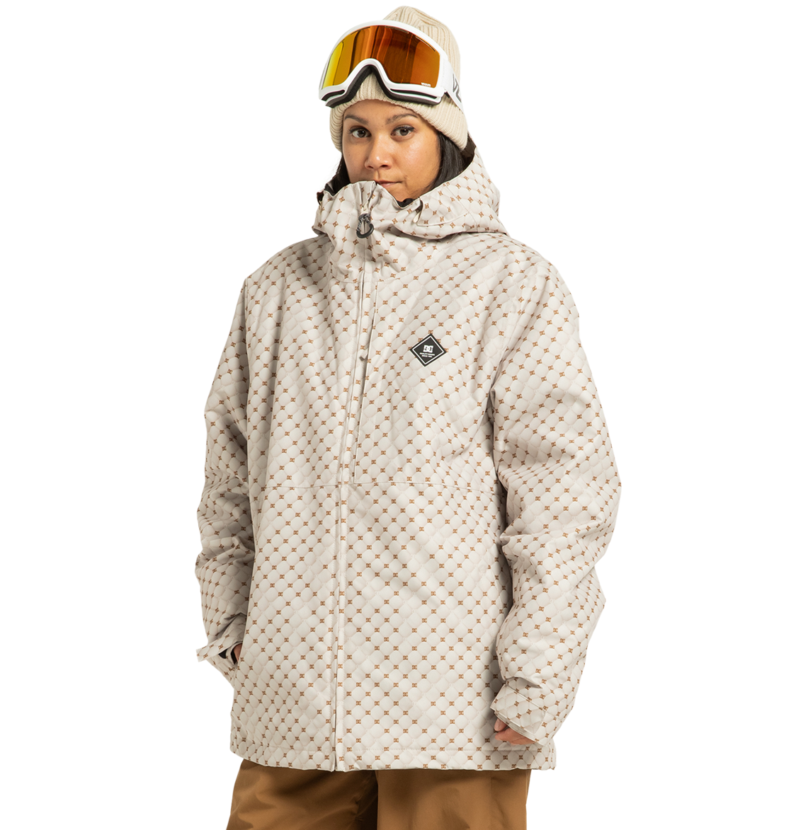 A person stands against a white background wearing the DC Shoes Unisex Basis Print Insulated Jacket—crafted from recycled polyester with a beige patterned design—paired with tan pants, a beige beanie, and ski goggles.