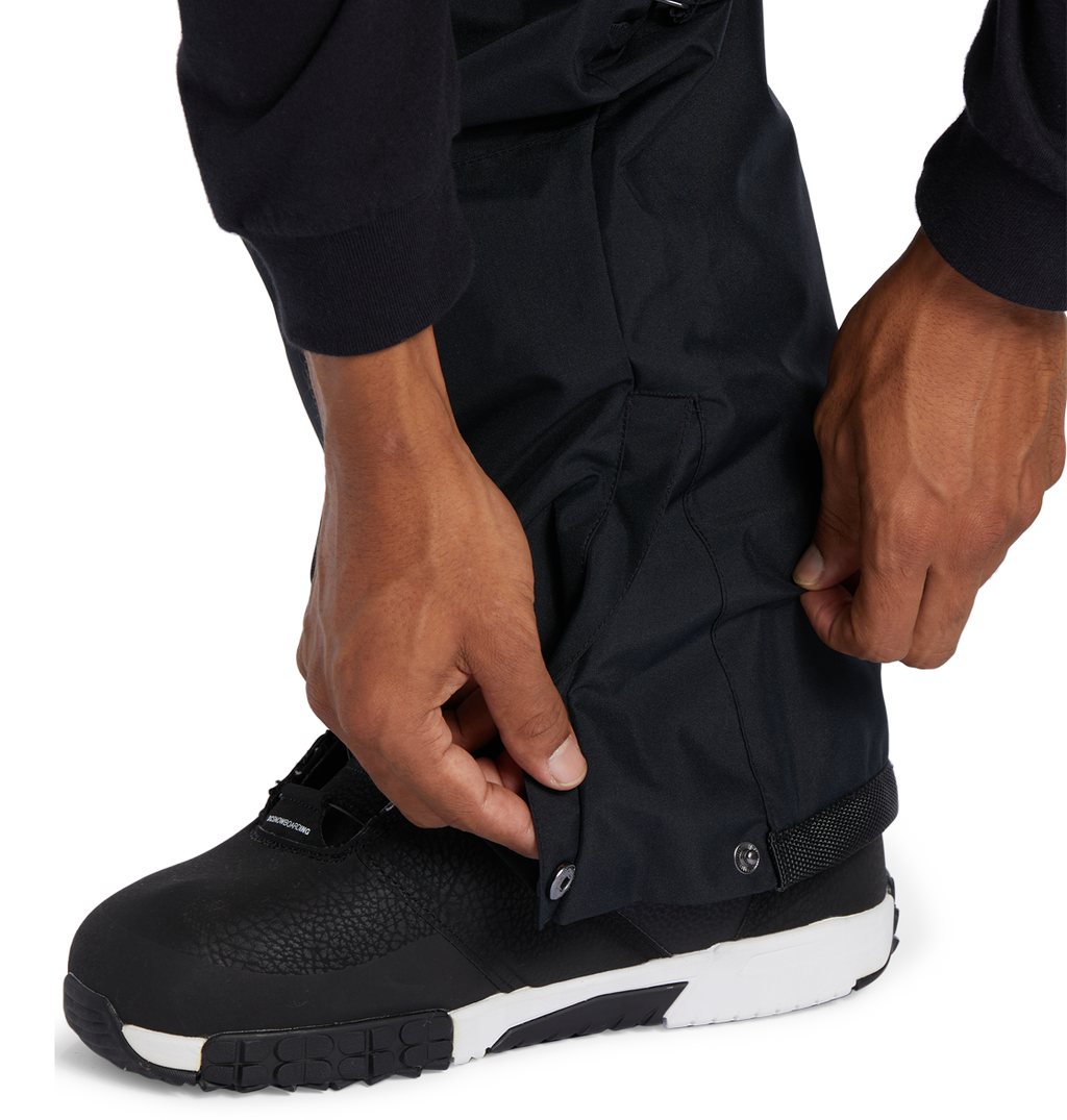 A person adjusts the snap button on the cuff of DC Shoes Men’s Banshee Insulated Pants, worn over black and white outdoor boots—an essential piece of winter gear.