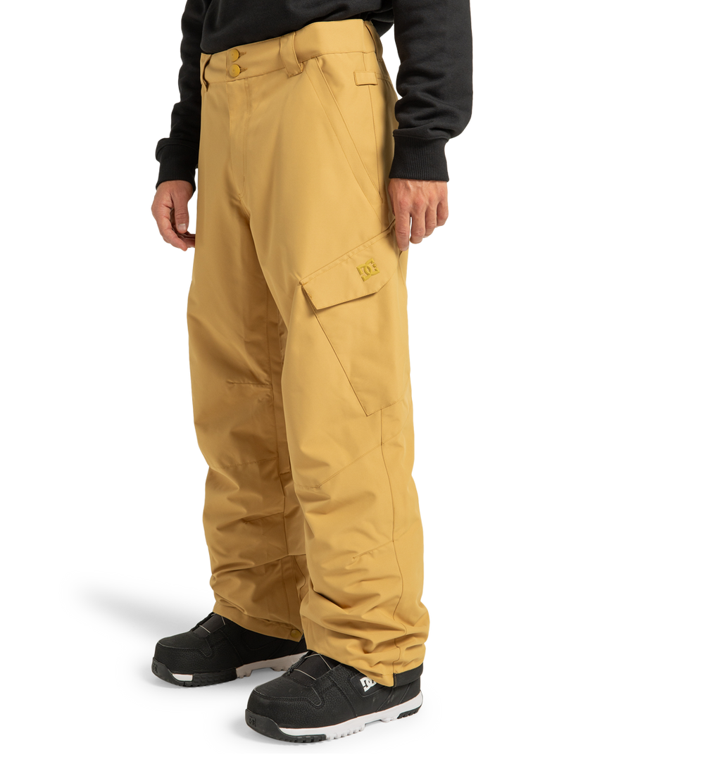 A person stands against a plain white background with their hands by their sides, wearing loose-fitting, tan DC Shoes Men’s Banshee Insulated Pants made from recycled polyester, paired with black sneakers and a black long-sleeve top.