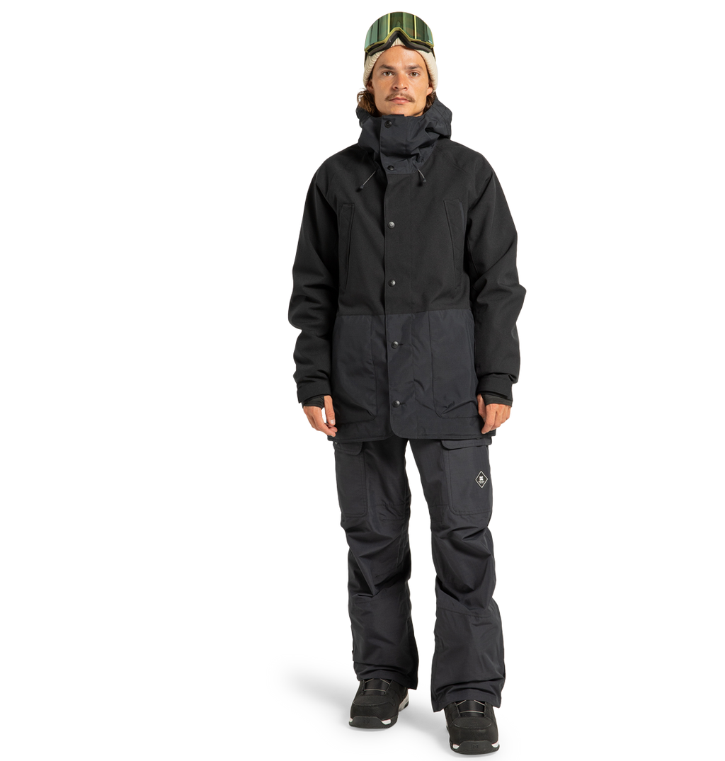 A person standing in snowboarding gear, featuring a black jacket, DC Shoes Men’s Code Shell Pants designed for eco-friendly performance, black boots, and green ski goggles resting on their head, against a plain white background.