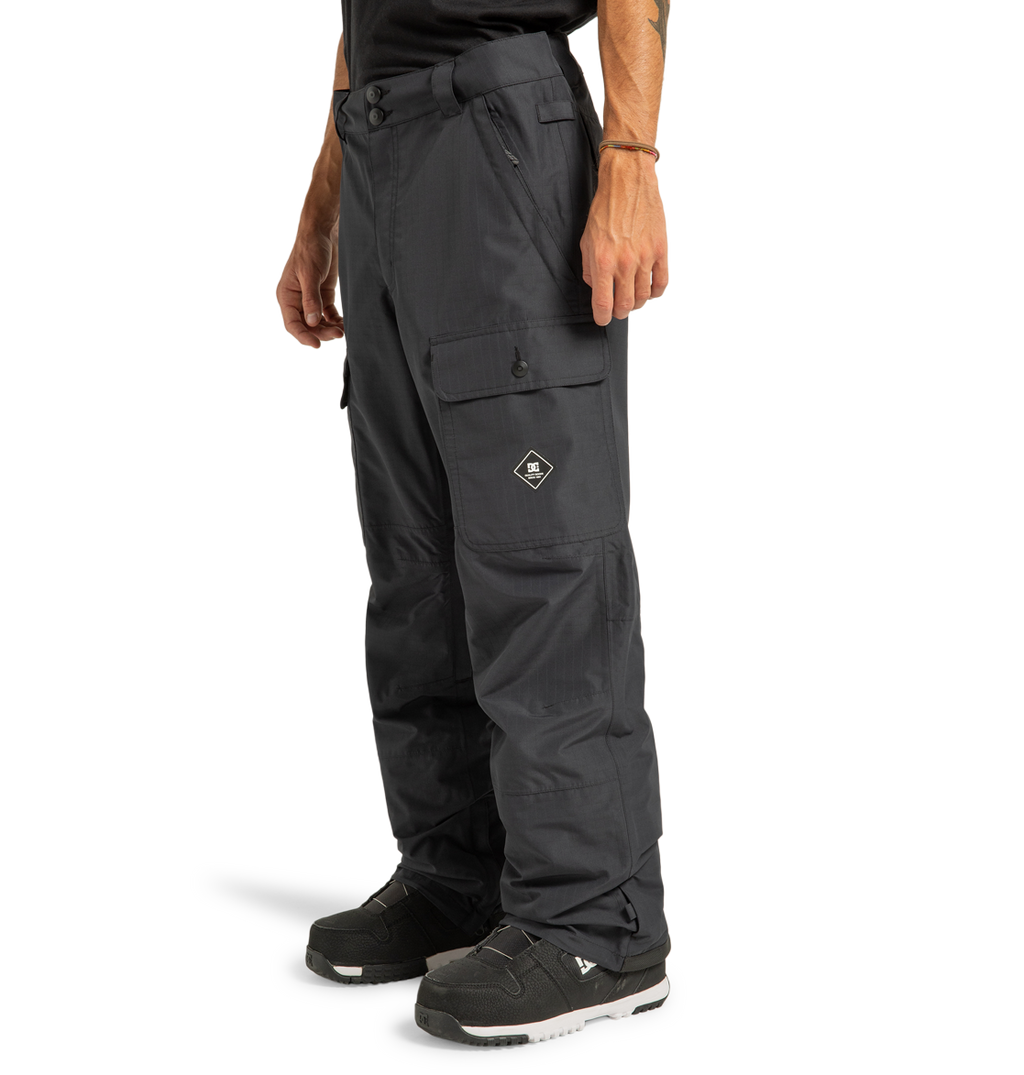 A person wearing DC Shoes Men’s Code Shell Pants in black, featuring eco-friendly materials and flap pockets, paired with black sneakers, stands against a plain white background.