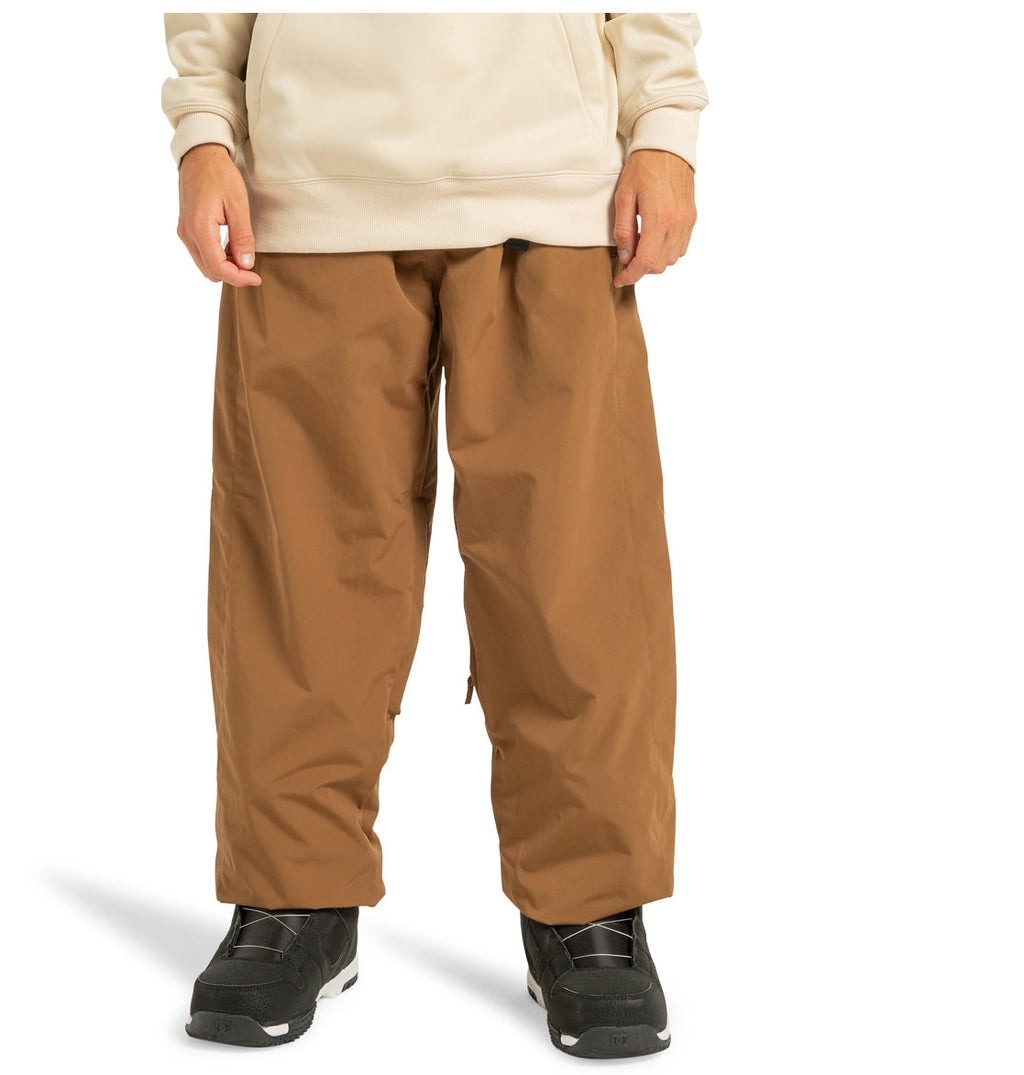 A person is standing against a plain white background, dressed in light brown DC Shoes Unisex Primo Shell Pants, a beige sweatshirt, and black sneakers. Renowned for their weather defense features, the Primo Shell Pants are a popular choice among outdoor enthusiasts. The image is cropped at the chest, leaving the person’s face out of view.