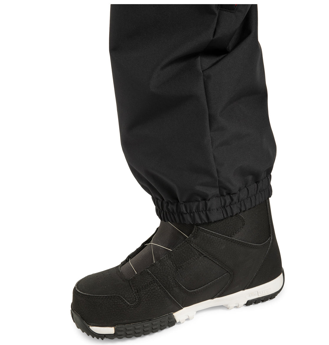 A close-up of a person wearing DC Shoes Unisex Primo Snow Pants, made from recycled polyester, paired with black snow boots featuring white soles, standing on a white background.