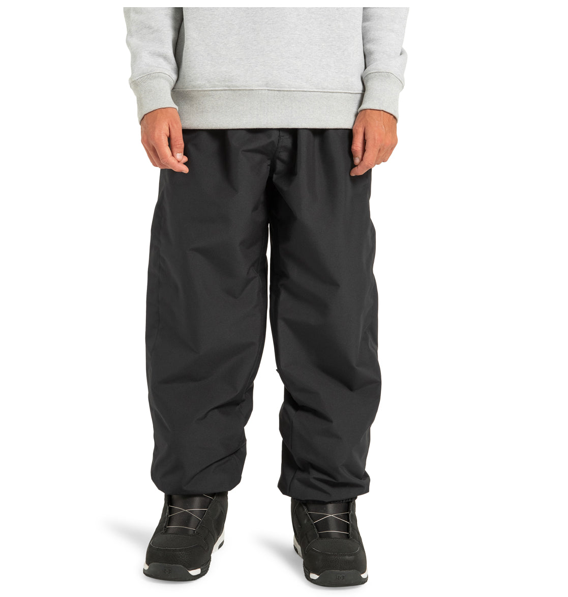 A person is wearing DC Shoes Unisex Primo Snow Pants in black, made from recycled polyester, along with a gray sweatshirt and black athletic shoes, standing against a plain white background.