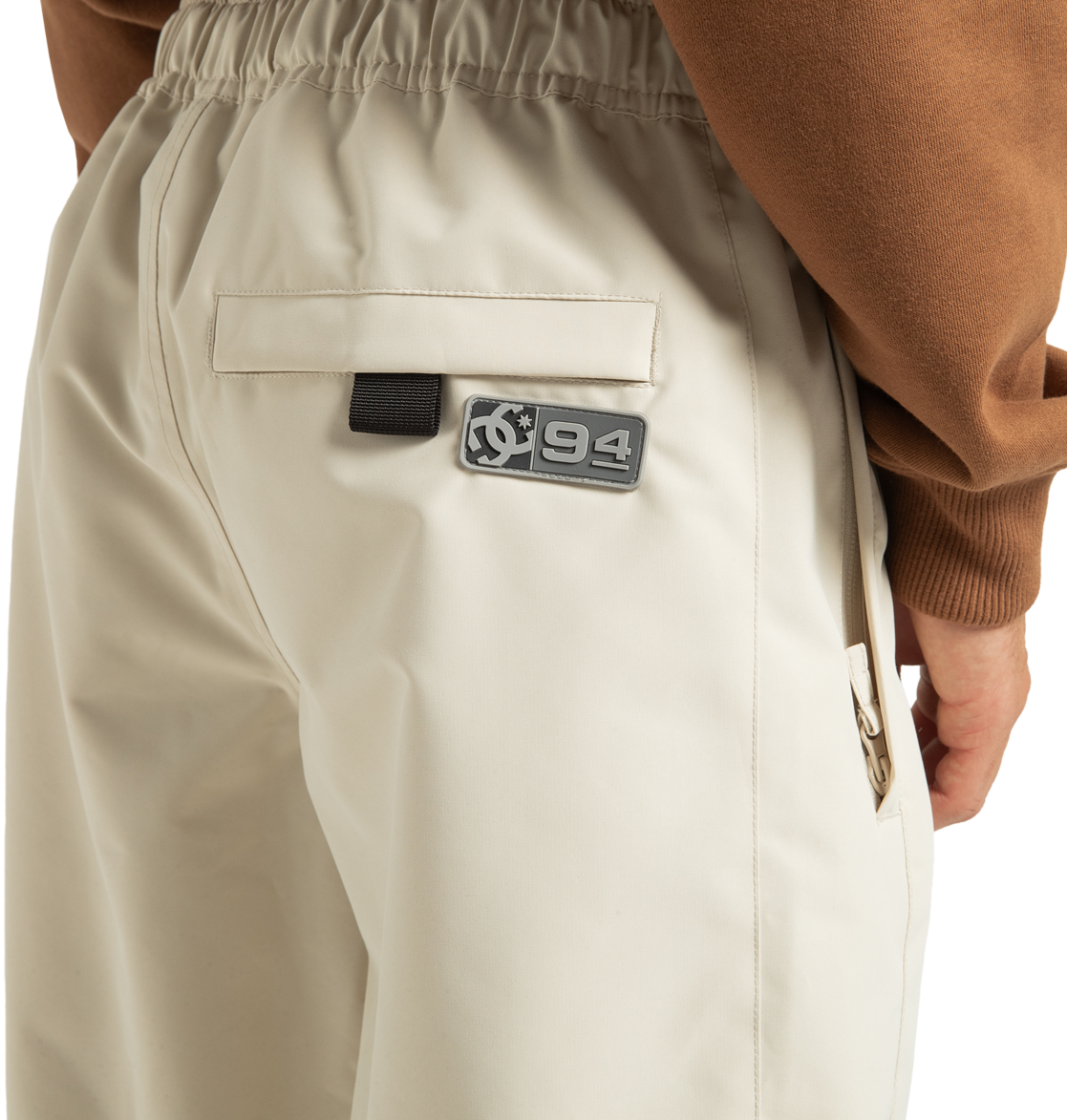 A close-up of a person wearing the DC Shoes Unisex Primo Snow Pants in beige, featuring an elastic waistband, a back pocket, and a patch reading “94” beside a circular logo. Made from recycled polyester, these waterproof snow pants are styled with a brown top.