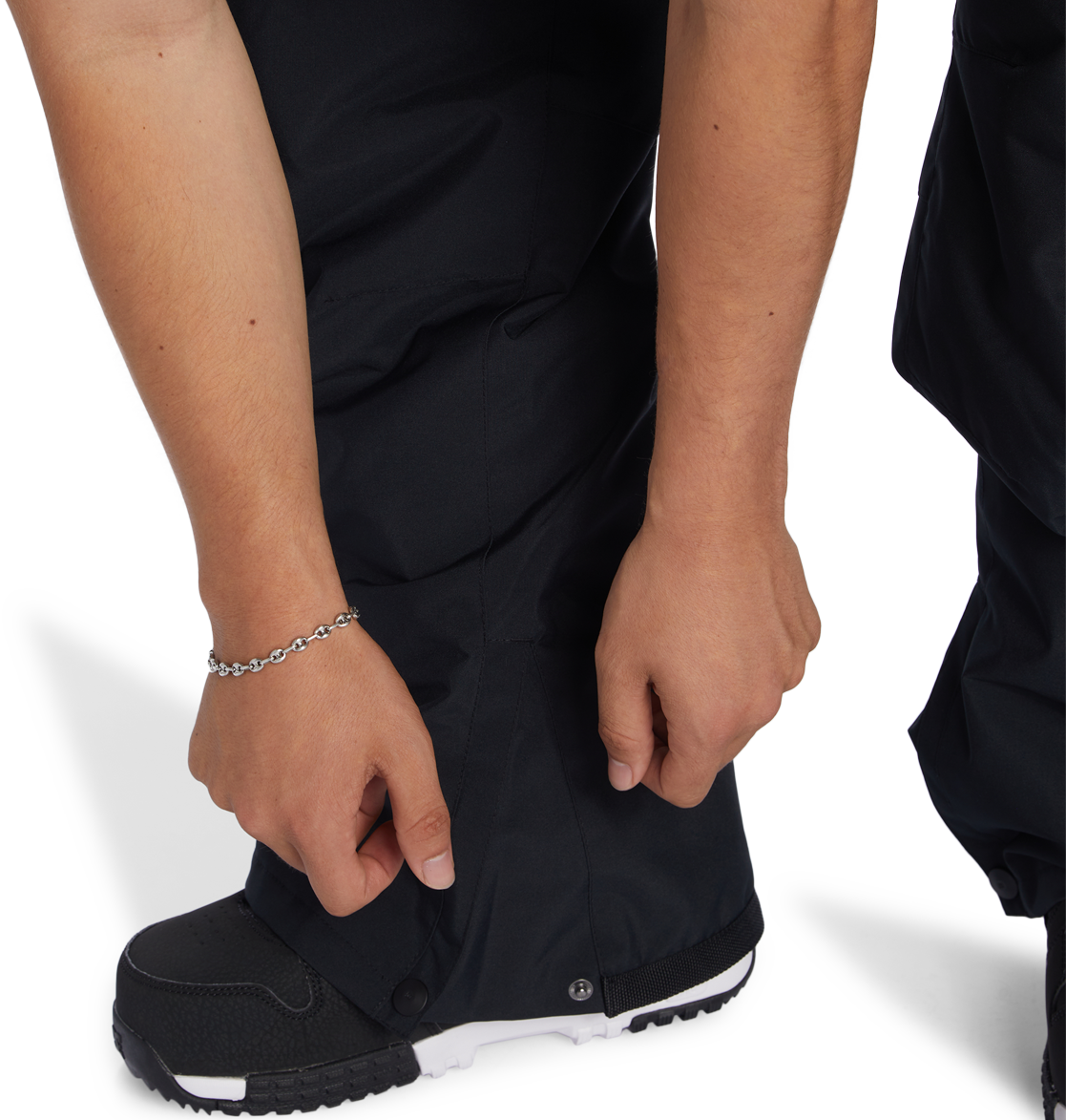 A person wearing DC Shoes Men’s Insulated Chino Pants in black and black-and-white shoes adjusts a snap at the ankle of their pants. Their left wrist has a silver chain bracelet, and the background is plain white.