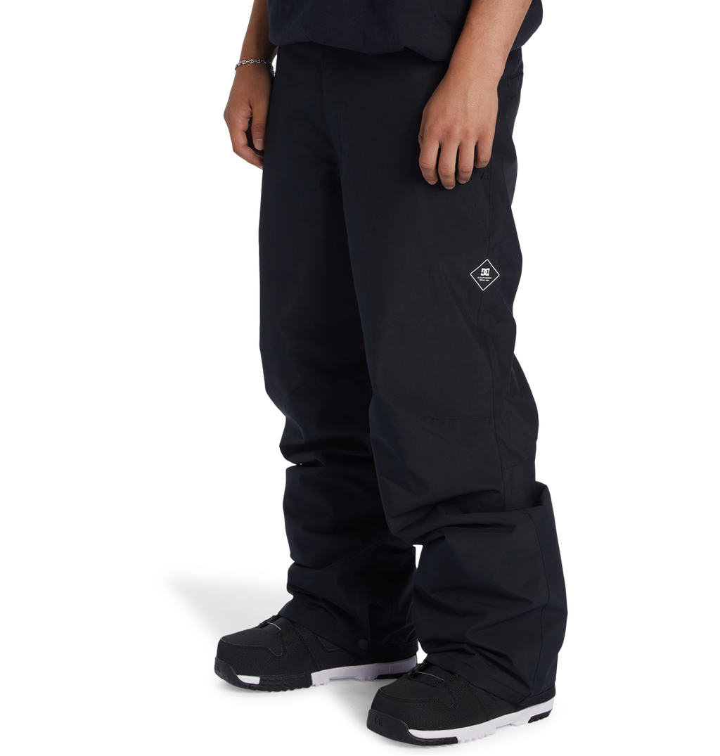 A person is shown from the waist down wearing DC Shoes Men’s Insulated Chino Pants in black, featuring a small white DC Shoes logo on the left thigh, paired with black sneakers with white soles and standing against a plain white background.