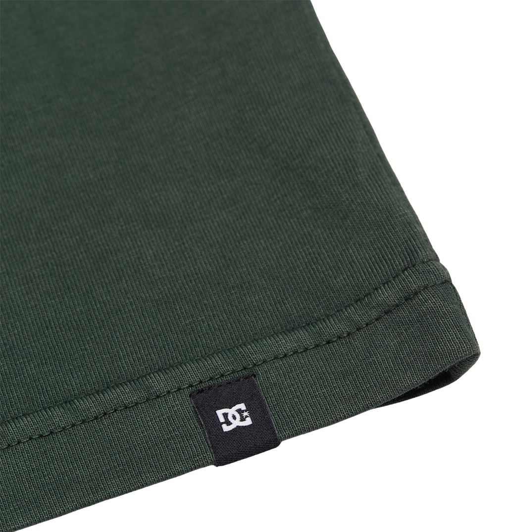 Close-up of the hem of the Men's DC Star Pigment Dye Tee by DC Shoes in dark green ribbed fabric, featuring a small black tag with a white DC logo sewn into the edge—offering understated branding reminiscent of classic DC artwork seen on graphic skate tees.