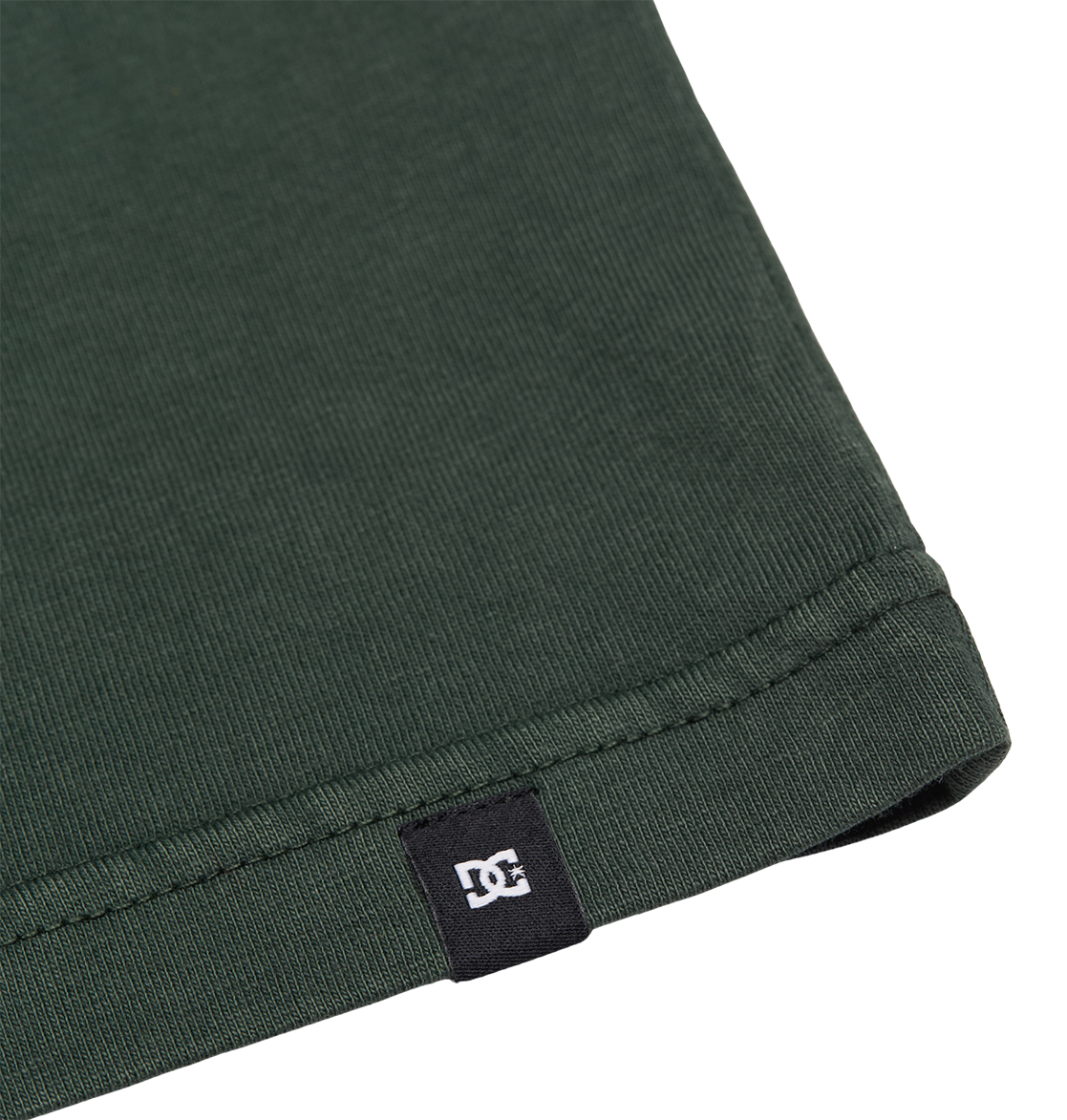 Close-up of the hem of the Men's DC Star Pigment Dye Tee by DC Shoes in dark green ribbed fabric, featuring a small black tag with a white DC logo sewn into the edge—offering understated branding reminiscent of classic DC artwork seen on graphic skate tees.