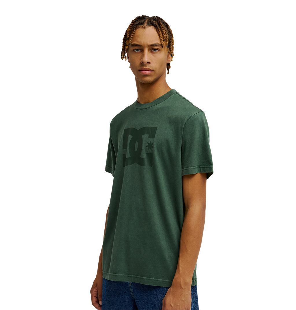 A person wearing the Men's DC Star Pigment Dye Tee by DC Shoes in dark green with a relaxed fit and blue jeans stands against a plain white background, looking straight ahead with a neutral expression.