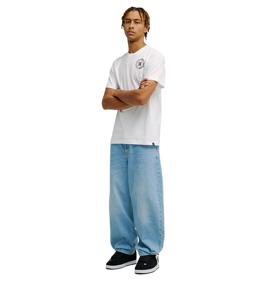 A person stands with arms crossed, wearing the Men's DC Corpo Tee by DC Shoes with a small circular logo on the chest, paired with light blue baggy jeans and black sneakers against a plain white background.