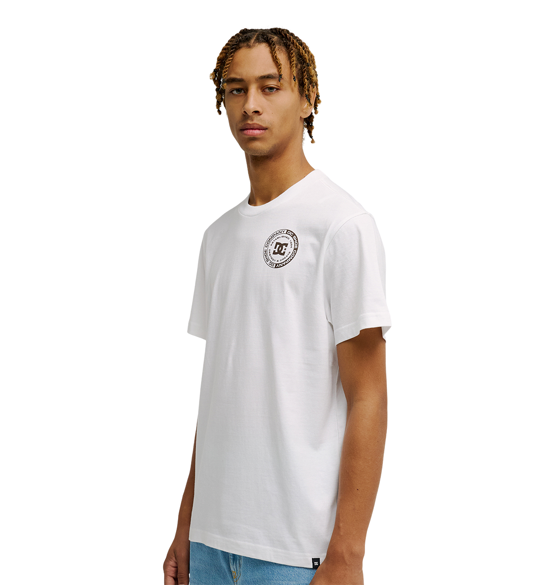 A young man with short, twisted hair models the Men's DC Corpo Tee by DC Shoes—a white relaxed fit t-shirt with a corporate logo—paired with light blue jeans. He stands against a plain white background, facing the camera.
